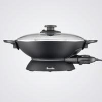 Black electric wok with glass lid on a light background, featuring the Breville brand.