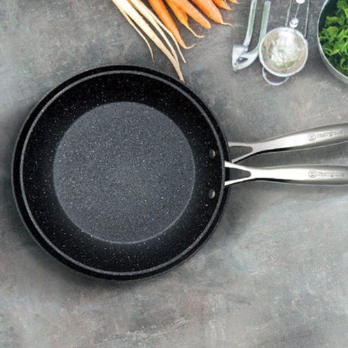 Black Westinghouse frying pans on a grey surface with carrots and kitchen utensils in the background