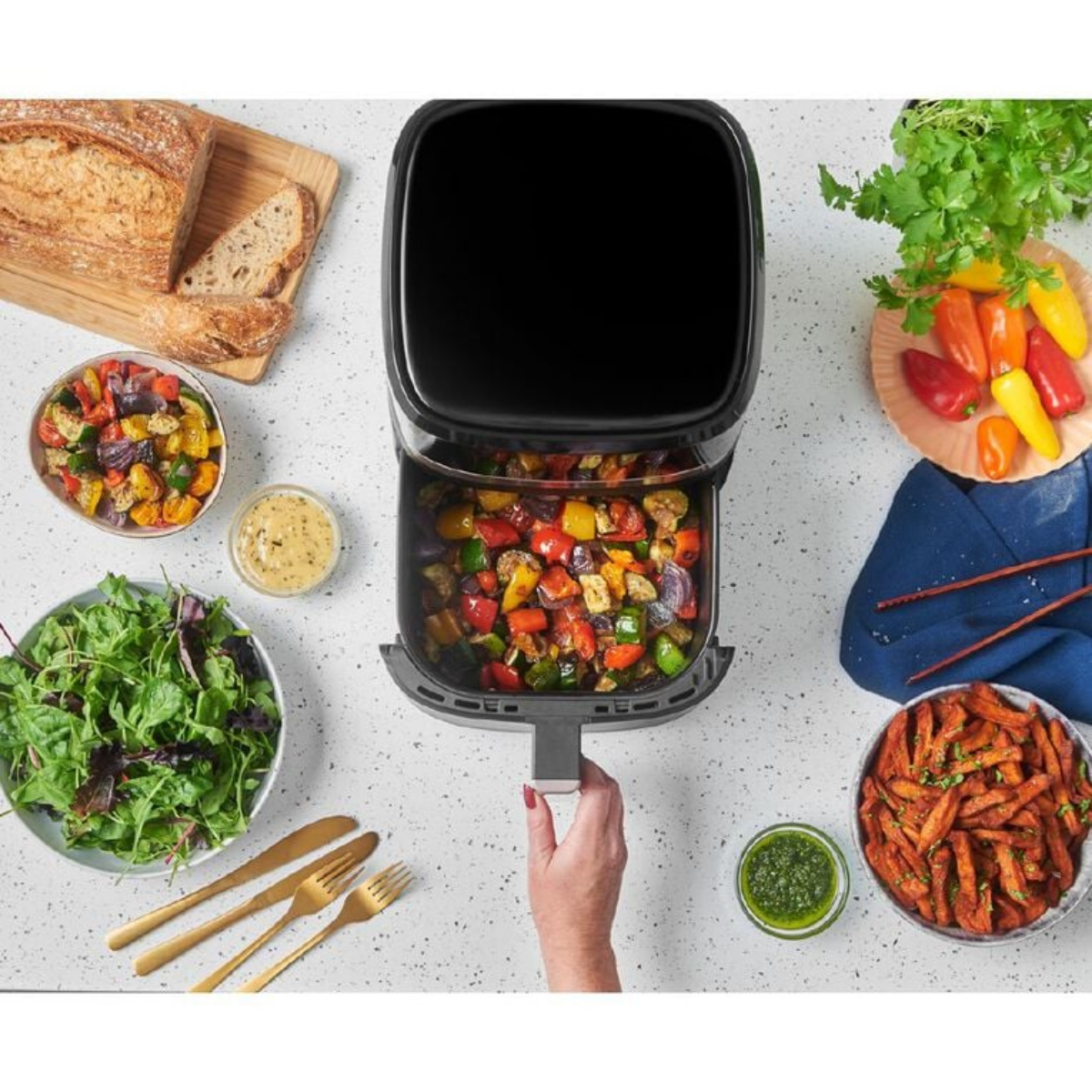 Black air fryer with roasted vegetables on a kitchen counter with various dishes and utensils.