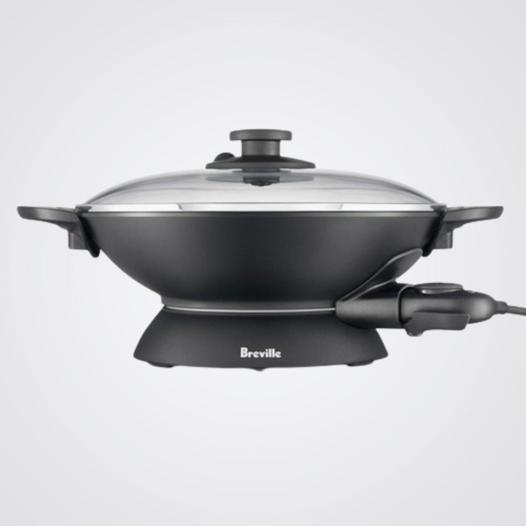 Black electric wok with glass lid on a light background, featuring the Breville brand.