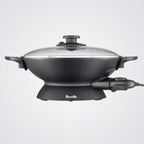 Black electric wok with glass lid on a light background, featuring the Breville brand.