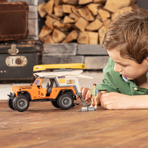 Child playing with a toy camper truck on a wooden floor with logs and trunks in the background.