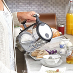 Person holding a transparent glass teapot with black lid over a breakfast table.